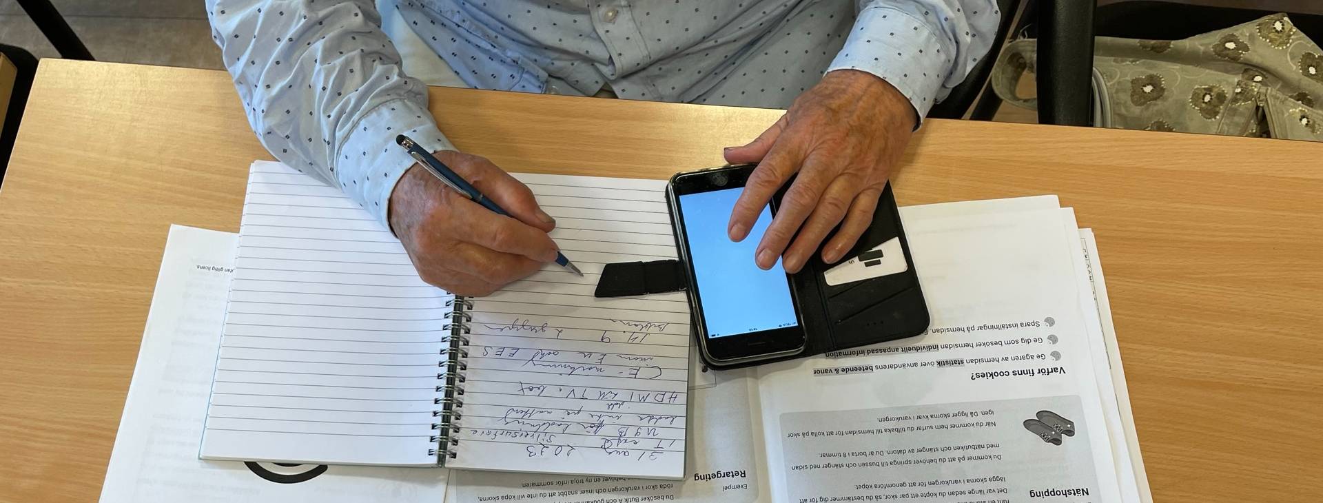 Händerna på en person som sitter vid ett skrivbord med en mobiltelefon och anteckningsblock.
