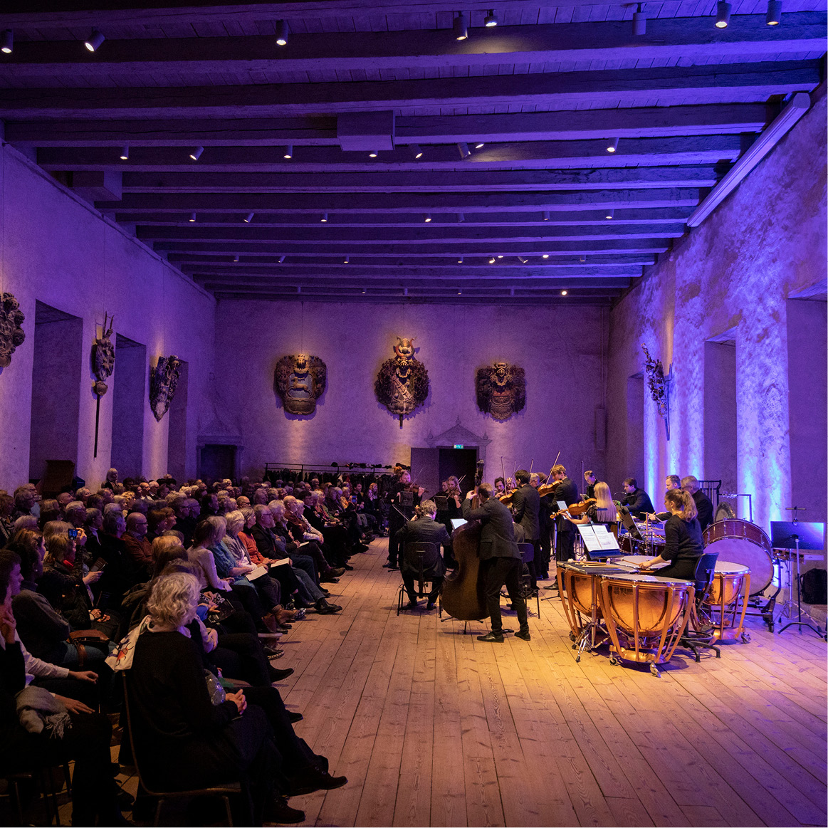 Vi ser en färgsatt sal i lila och där syns en orkester med fioler och violas, samt cellos.