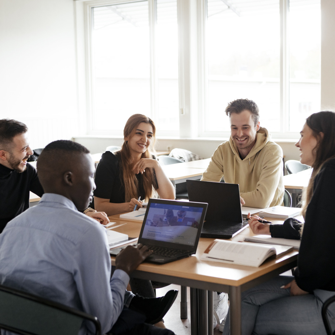 Fem personer sitter i ett klassrum vid ett bord och verkar glada. 