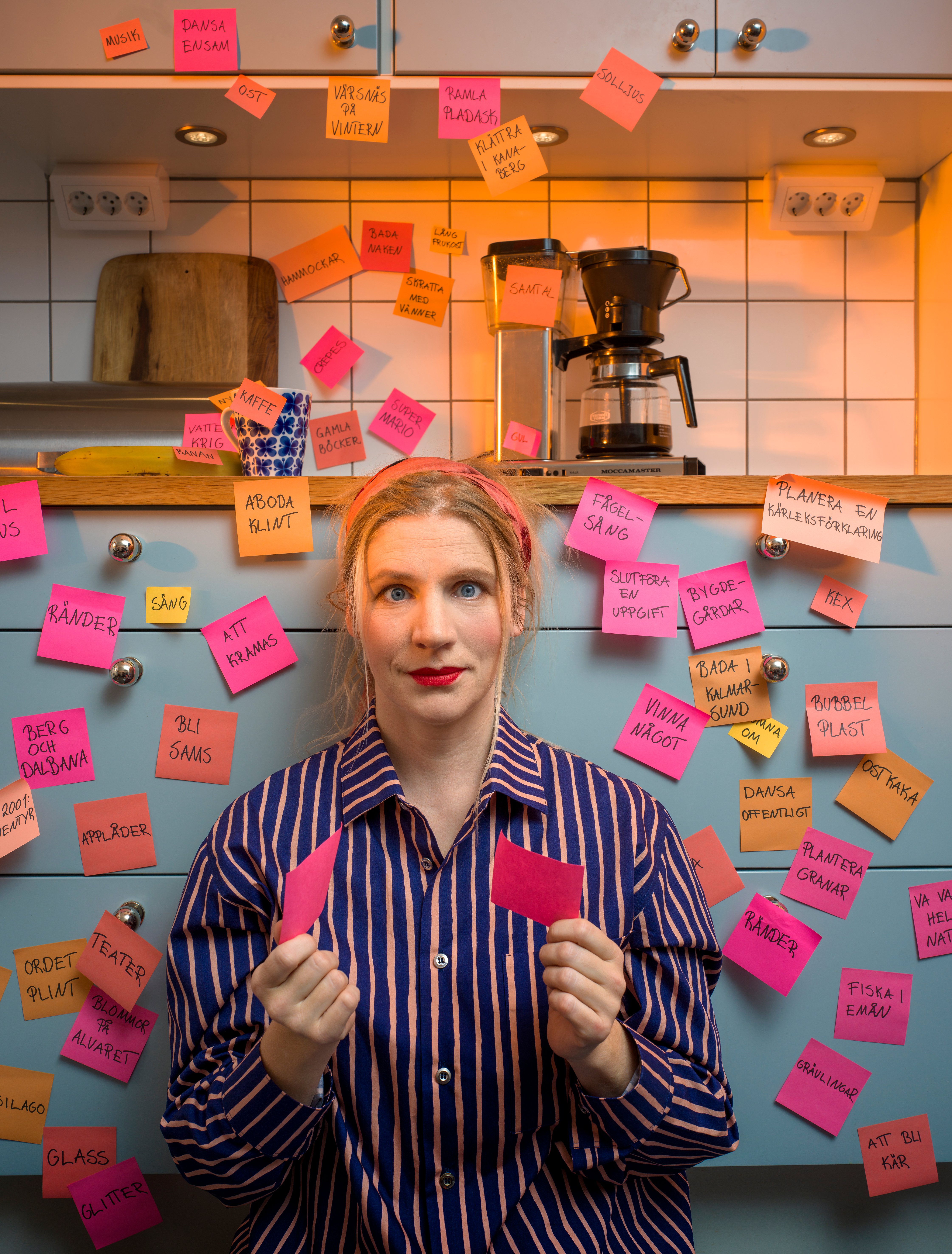 Bildtolkning: En kvinna sitter på golvet i ett kök omgiven av post-it-lappar. Hon har också post-it-lappar i händerna.