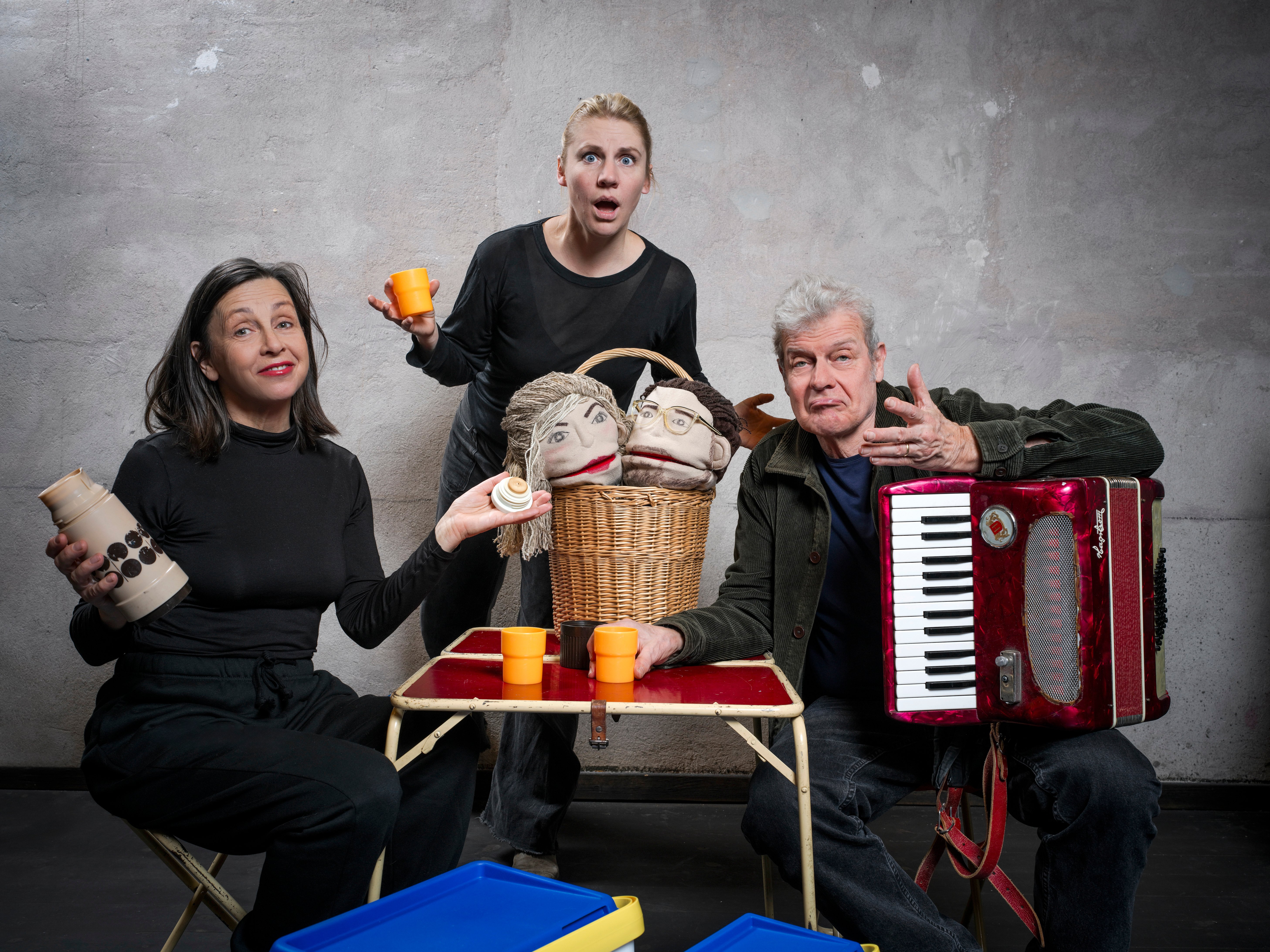 Bildtolkning: Tre personer sitter vid ett campingbord för en fikastund. EN har ett dragspel i famnen. Alla tre har ett förvånat, undrande minspel och har händerna i en frågande gest. 