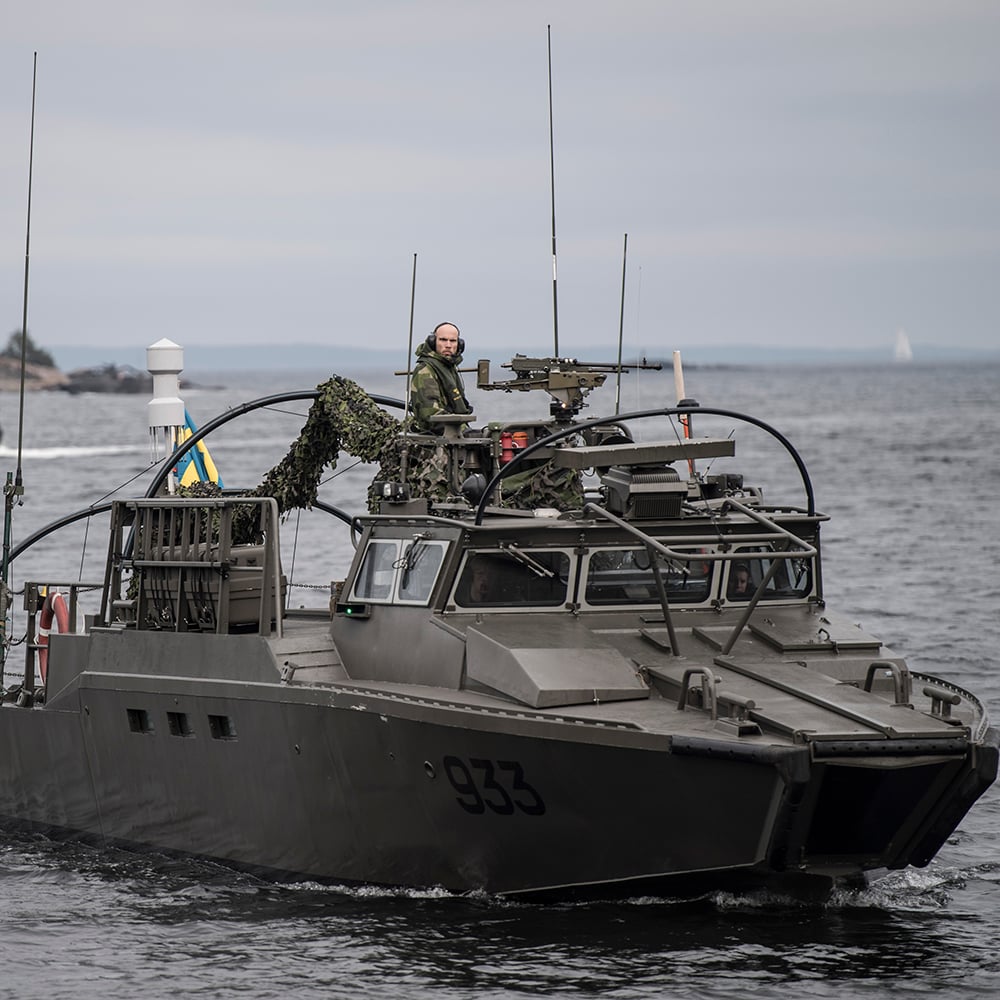 pressbild från Försvarmakten på en av marinens båtar med en soldat som tittar in i kameran och flera soldater i förarhytten