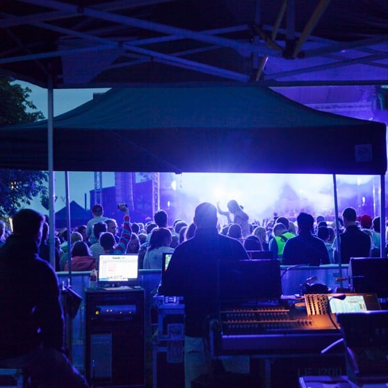 Personer vid mixerbord och publik framför en scen med en artist. 