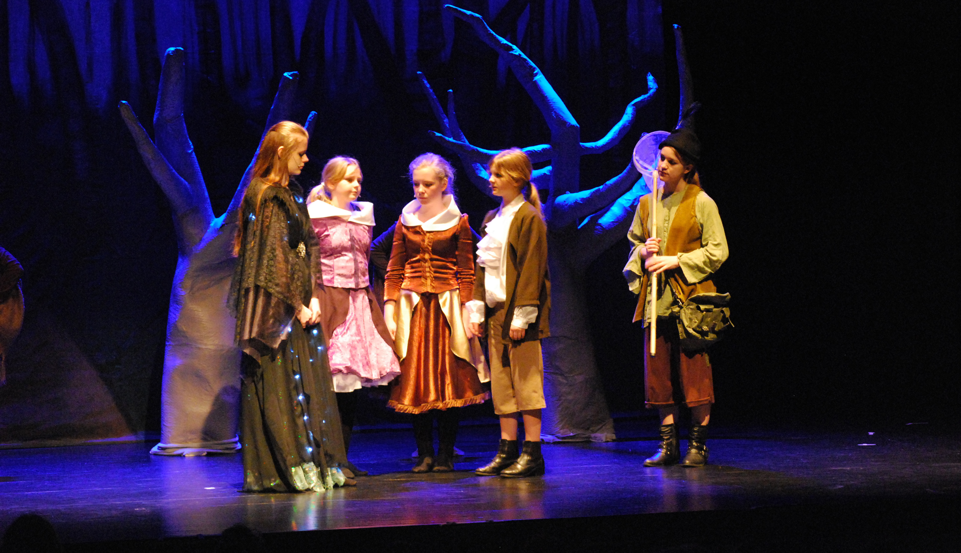 Dramaelever står på scenen i Bryggaren, en scen ur "Trollflöjten" med mörka träd i bakgrunden.